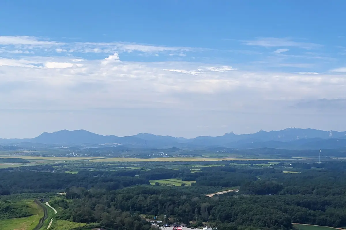 A wide view north from Dora Observatory over green forest and farmland toward a line of blue hills, with a flag mast on the right and a large cloudy sky above