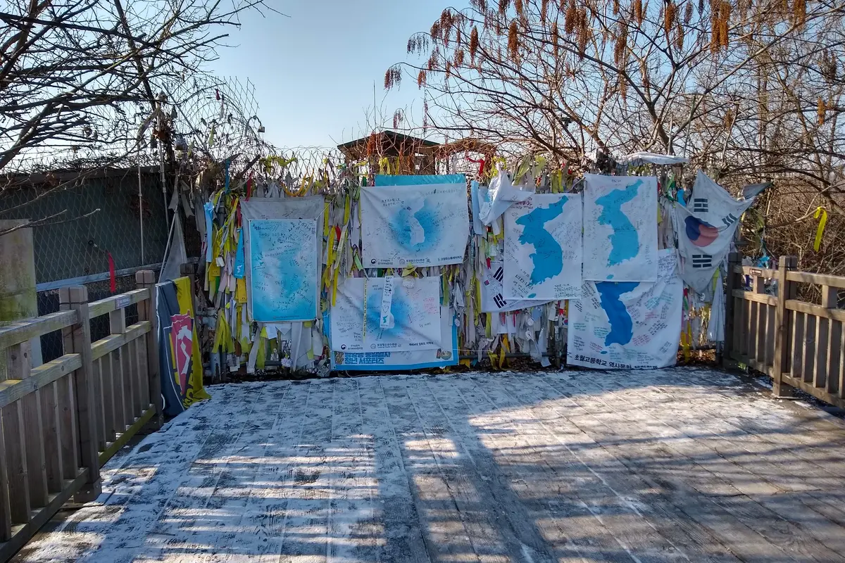 Ribbons, hand-drawn maps of Korea and flags tied to a wire fence beside a wooden walkway at Freedom Bridge, with bare trees and a blue sky behind